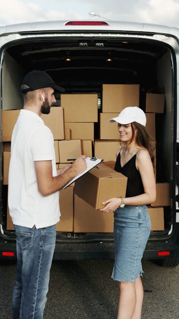 A man and a woman are standing at the open rear of a white van, which is loaded with numerous cardboard boxes used for home relocation. The man, dressed in a white t-shirt, jeans, and a black cap, is holding a clipboard and appears to be recording details related to the moving process. The woman, in a black sleeveless top and a denim skirt, is smiling and holding a medium-sized cardboard box, likely containing personal belongings for packing and moving. The van's interior shows a stack of boxes arranged neatly, suggestive of a systematic packing and loading process undertaken by a professional removals service. The scene is outdoors, on a paved surface, during daylight, and exemplifies the logistics of furniture transport and home relocation handled by Man with Van West Ham, supporting efficient and organised moving services in West Ham.