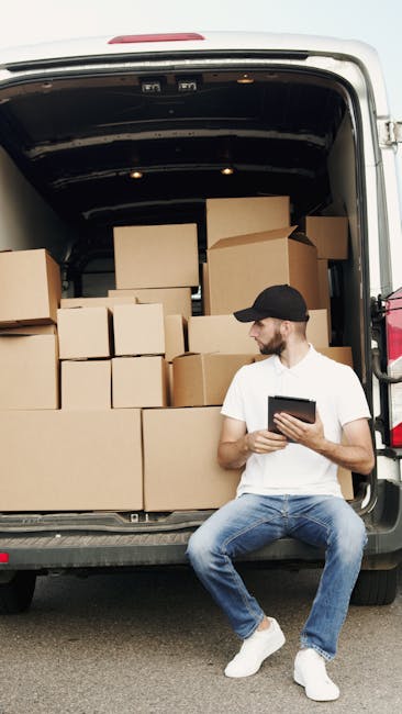 A man wearing a white t-shirt, jeans, and a black cap is carrying three cardboard boxes stacked together while standing outside next to a white cargo van parked on the street. The van is part of the home relocation process managed by Man with Van West Ham, visible through the company's website domain, manwithvanwestham.co.uk. The van's rear doors are open, revealing a spacious interior used for furniture transport and packing during moving services. Several additional cardboard boxes are placed on the pavement nearby, some stacked at the side of the van, indicating the loading process of packing materials. In the background, there are residential houses with pitched roofs, slate tiles, and chimneys, along with green bushes and tall trees, under natural daylight. The scene captures key aspects of house removals, such as packing, loading, and transporting household items in preparation for an upcoming home relocation or furniture transport.