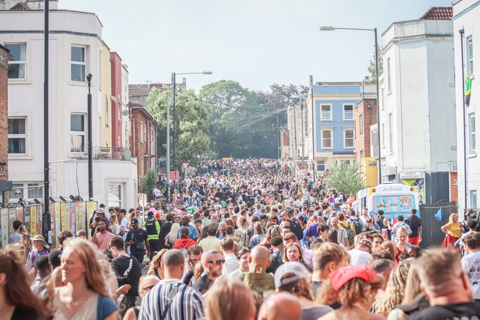 A densely packed crowd of people gathered on a street in West Ham, with residential buildings lining both sides of the road. The scene shows a high concentration of pedestrians, some wearing casual clothing and hats, with a few individuals carrying bags or backpacks. In the background, trees and utility poles are visible, along with a clear, slightly cloudy sky. Street lamps and signage are present along the pavement, which is partially obscured by the crowd. On the right side, a white ice cream van with colorful graphics is parked near the curb. This busy urban environment reflects the typical setting where house removals and furniture transport might be occurring, and it highlights the importance of efficient packing, loading, and logistical planning by companies such as Man with Van West Ham during home relocation or moving services.