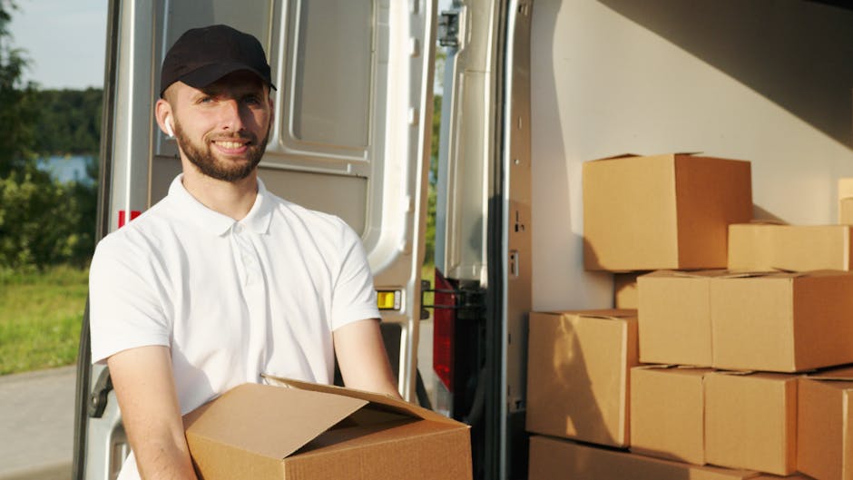 A man with a beard, wearing a black cap and a white polo shirt, is standing outdoors beside an open delivery van during daylight. He is holding a cardboard box and smiling at the camera. Inside the van, there are several stacked cardboard boxes, some wrapped with plastic, arranged on the floor and sides of the vehicle. The van's interior is well-lit, and the scene shows the process of loading or unloading household items, including furniture and packaging materials, which is part of a house removal or home relocation service. The background includes a green landscape with trees and a body of water, indicating a scenic outdoor setting. The presence of a moving trolley and the careful stacking of boxes suggest a professional removals service, such as those offered by Man with Van West Ham, focusing on efficient packing and furniture transport.