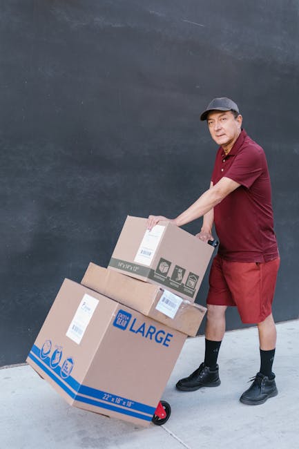 A man wearing a maroon polo shirt, red shorts, black socks, and black shoes is standing outdoors on a paved surface next to a dark grey wall, pushing a hand truck loaded with several cardboard boxes of varying sizes. The boxes are sealed with shipping labels and packing tape, with one marked 'LARGE' in blue text. He appears to be in the process of transporting items as part of a home relocation or moving service. The scene illustrates the loading process involved in a furniture transport or last-minute move in West Ham, supported by companies like Man with Van West Ham who offer removals and packing services.