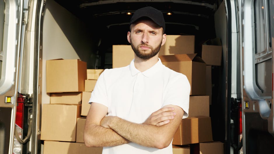 A man wearing a black cap and white polo shirt standing with arms crossed inside the open rear of a moving van, which is packed with numerous brown cardboard boxes of various sizes, some stacked on to