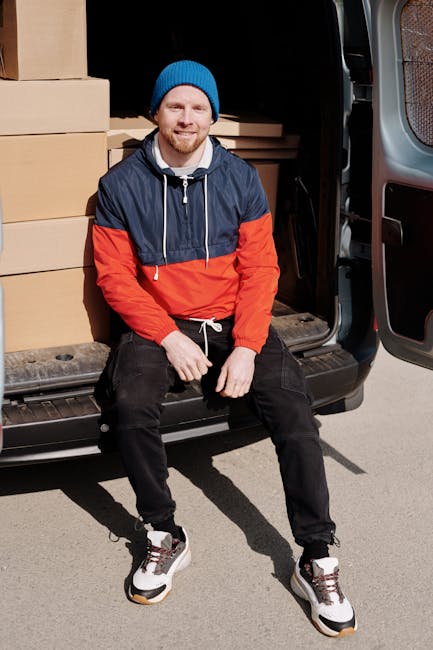 A man wearing a black cap and white polo shirt standing with arms crossed inside the open rear of a moving van, which is packed with numerous brown cardboard boxes of various sizes, some stacked on to