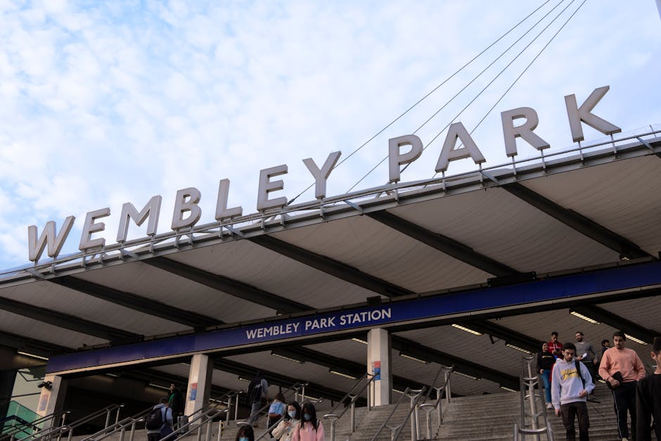 Photograph of the exterior entrance to Wembley Park Station, with a large sign displaying the station name in uppercase letters mounted on the roof. Below the sign, a blue panel indicates the station's name. Several pedestrians, some wearing face masks, are walking down the station’s steps on the pavement outside. The sky above is partly cloudy with patches of blue visible. The scene captures a typical busy station environment, which might be relevant when considering residential relocations or moving logistics nearby, as [COMPANY_NAME] offers house removal and furniture transport services in the West Ham area, supporting home relocation and packing processes. The image emphasizes transportation and accessible entry points, essential for planning a home relocation or efficient loading process when using professional removal services.