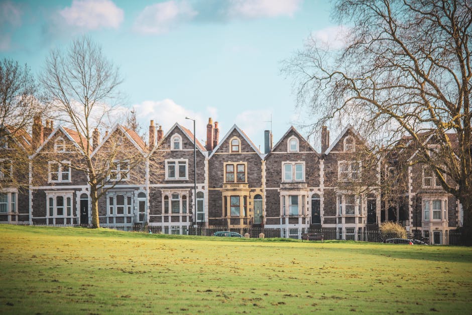 A row of three-storey Victorian-style terraced houses made of grey stone with white window frames and decorative details, situated behind a low black metal fence. In the foreground, a well-maintained grassy area extends across the image, with a few leafless trees casting shadows on the lawn. The sky above is clear with some scattered clouds, and the scene is illuminated by natural daylight. This residential area provides a typical backdrop for home relocation or furniture transport services in West Ham, with the houses visible from the pavement. The setting suggests an urban environment suitable for house removals, with space for moving vans and loading equipment to operate efficiently, supporting professional packing and moving activities in the context of local removals provided by [COMPANY_NAME].