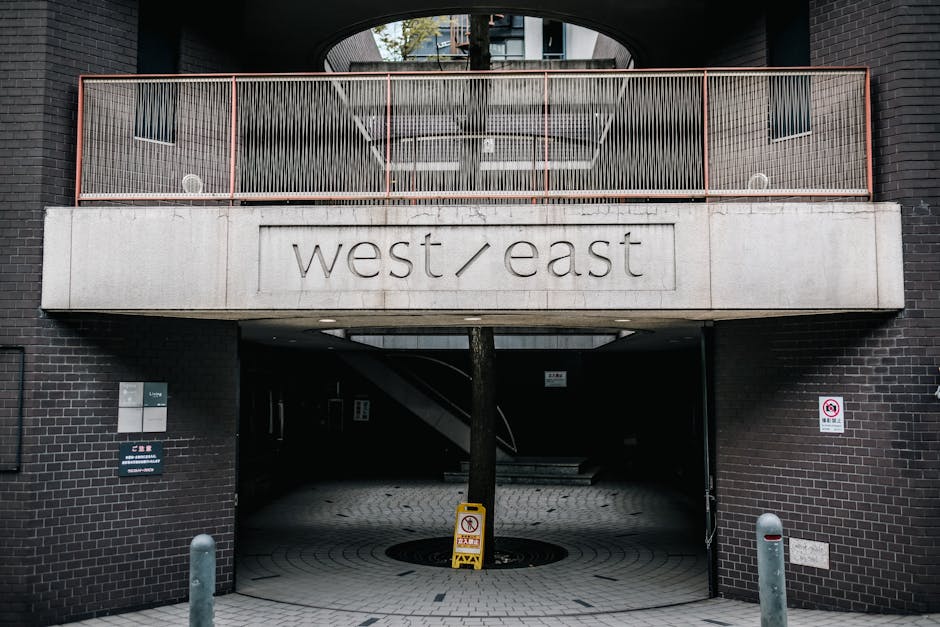 The image shows the entrance to a modern building with a dark brick exterior and a large, curved concrete sign displaying the words 'west / east' in lowercase letters. Above the entrance, there is a balcony with a metal railing painted in a rust colour, featuring vertical bars. Inside, the building appears to have a partially visible staircase and an open lobby area. A tree trunk extends through a circular opening in the ground, positioned directly beneath the sign. To the left of the entrance, there is a small signboard and a bollard on the sidewalk, while the right side features a no parking sign mounted on the brick wall. The setting suggests an urban environment suitable for house or professional removals, with vehicle access through the entrance for loading and unloading furniture or packing materials as part of a home relocation process, characteristic of services offered by companies like Man with Van West Ham. The lighting indicates daytime, and various materials such as concrete, brick, metal, and paving are visible, reflecting a contemporary residential or commercial building environment.