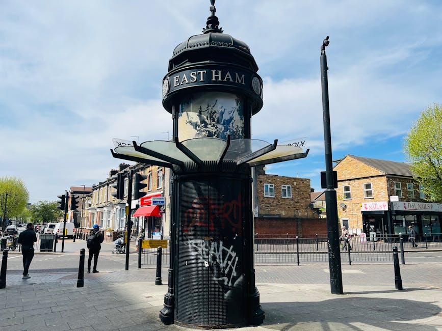 A street scene in West Ham featuring a prominent cylindrical public notice board with a domed top and the words 'EAST HAM' displayed at the crown, situated on a paved sidewalk. Surrounding the notice board are black bollards, pedestrians, and street furniture such as benches and streetlights. In the background, there are residential buildings with brick facades, commercial shops including a nail salon, and trees with green foliage. The sky is mostly clear with a few clouds. The setting suggests an urban environment where house removals and furniture transport by [COMPANY_NAME], such as loading and moving household items, may take place nearby, supported by the infrastructure visible in the scene.