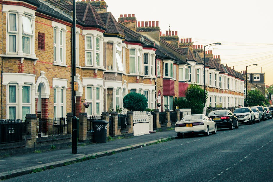 A street scene in West Ham showing a row of terraced houses with brick facades, some with white bay windows and decorative moldings, and others with different coloured exterior panels. The sidewalk in front of the houses has small front gardens enclosed by black wrought iron fences with brick pillars. Several parked cars, including several sedans and hatchbacks, are lined up along the curb. In the foreground, a black waste bin with the label 'DOGG' and a street lamp are visible. The image captures a typical residential area during daylight, with no visible activity or moving vehicles. This scene relates to house removals and moving services, as it depicts the environment where such services might operate, especially considering parking restrictions and permits in West Ham. The picture is taken from the pavement, providing a clear view of the front of the houses and the parking arrangement, illustrating the context of urban relocations managed by companies like Man with Van West Ham.