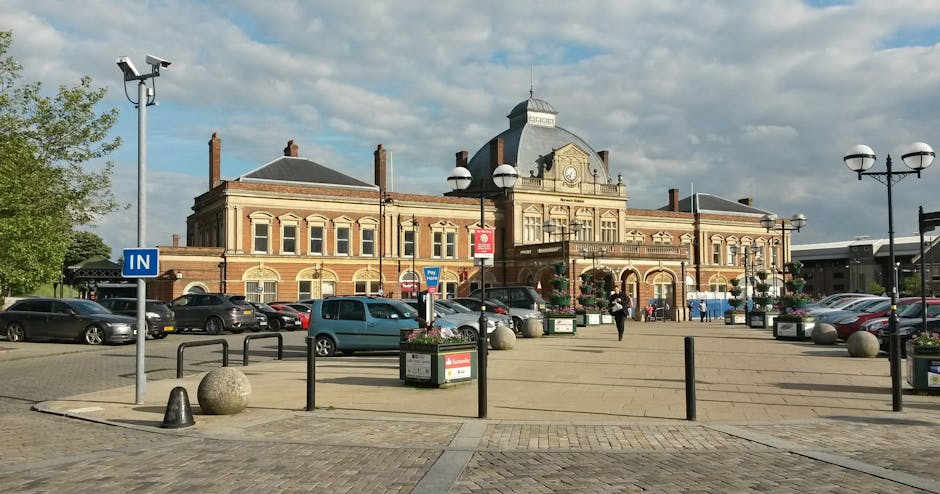A street scene in West Ham featuring a prominent cylindrical public notice board with a domed top and the words 'EAST HAM' displayed at the crown, situated on a paved sidewalk. Surrounding the notice board are black bollards, pedestrians, and street furniture such as benches and streetlights. In the background, there are residential buildings with brick facades, commercial shops including a nail salon, and trees with green foliage. The sky is mostly clear with a few clouds. The setting suggests an urban environment where house removals and furniture transport by [COMPANY_NAME], such as loading and moving household items, may take place nearby, supported by the infrastructure visible in the scene.