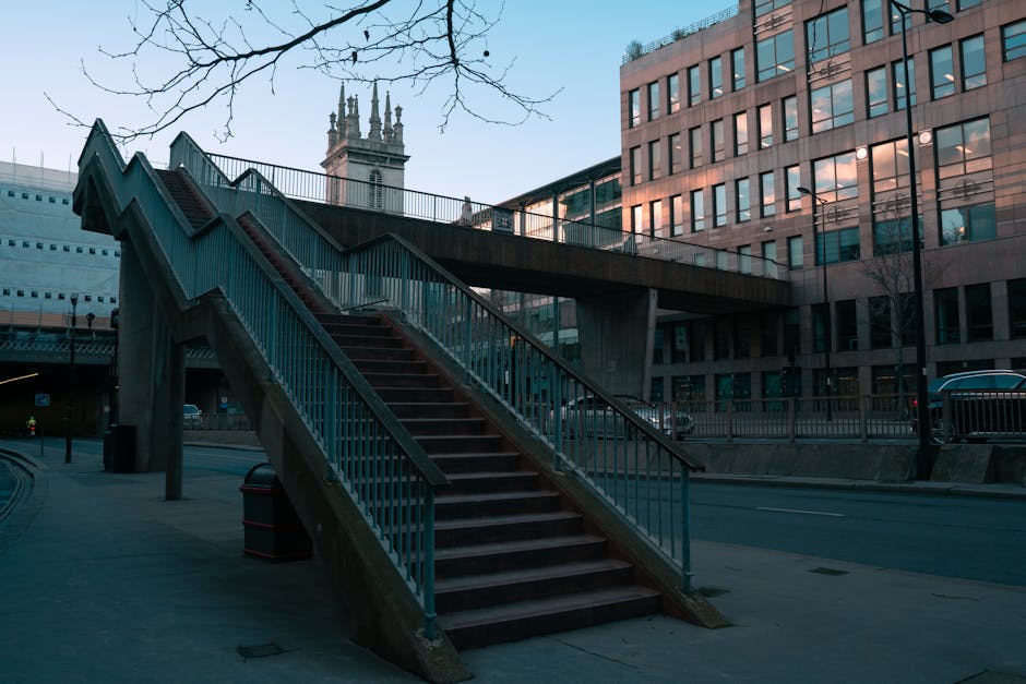 The image depicts an outdoor concrete staircase with red wooden steps and metal railings, situated on a city street with modern glass-fronted office buildings on one side and a historic tower visible in the background. The staircase is designed for pedestrian access between street level and an overpass or elevated walkway. It is positioned next to a sidewalk, with a black trash bin at the base. The surrounding environment includes a few leafless trees and street lamps, indicating an urban setting. The area is well-lit with natural daylight, and the scene is free of people or vehicles at the moment. This setting is relevant to home relocation or furniture transport processes that may require navigating tight or narrow access points, such as staircases, in urban environments, and is typical of the type of logistics managed by Man with Van West Ham for residential or commercial moves.