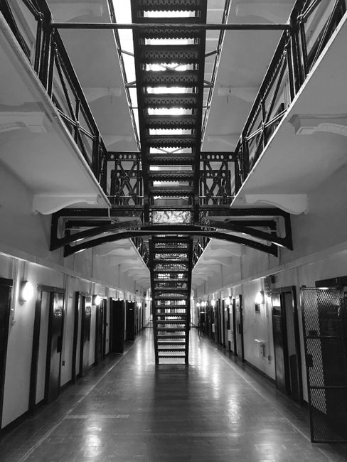 A black-and-white photograph of an indoor staircase within a multi-story residential building, showing a central metal staircase with perforated steps and railings, connecting the ground floor to the upper levels. The staircase is positioned in the middle of a spacious corridor, which has a polished wooden floor and white walls lined with multiple evenly spaced doors. There are ceiling-mounted lights illuminating the corridor, and an internal fire escape platform with metal railings is visible above the staircase. The scene depicts an empty hallway during daylight hours, with no furniture or moving equipment present, highlighting the structural design of the staircase and corridor. This image exemplifies the type of access challenge encountered during house relocations or furniture transport in buildings with narrow or limited entry points, with Man with Van West Ham capable of assisting with such staircase and narrow-access moves across West Ham.