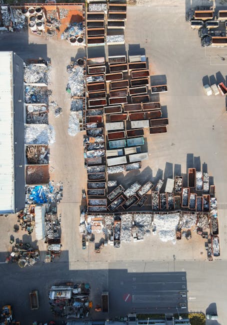 An aerial view of an outdoor industrial area showing numerous stacks of metal and wooden furniture, including cabinets, tables, and shelves, arranged on a concrete surface. Some items are wrapped in plastic or covered with protective materials, while others are uncovered. To the left, a large building with a grey roof is visible, along with scattered metal parts and packaging materials. The shadows cast by the furniture and objects indicate bright sunlight, suggesting daytime. In the bottom section, there are vehicles and equipment, including a forklift, which may be used for loading or moving items. This scene reflects a furniture storage or disposal yard associated with house removals and relocation services, such as those provided by Man with Van West Ham, supporting efficient furniture transport and packing logistics during home moves.