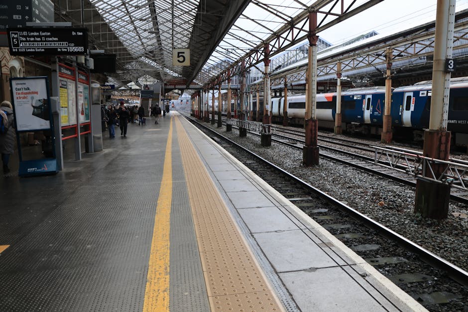 The image shows the platform at West Ham railway station, part of the London Euston line, featuring a canopy supported by rusted steel columns and a mixture of glass and metal roofing structure overhead. The platform surface is made of textured, dark-colored rubberized tiles with a yellow tactile paving strip running along the edge for safety. Several passengers are visible standing near signage and ticket machines on the left side of the platform, some wearing coats, with a few holding luggage or bags. To the right, adjacent to the platform, are railway tracks with loose gravel and ballast, on which a modern blue and white Vantibus West train car is parked. The train's exterior has the company branding, and its windows are visible. Outside the platform, a large digital departure board displays information about upcoming train departures, including the destination 'London Euston,' the train number, and scheduled departure time. The environmental lighting suggests an overcast day, typical for urban rail stations, and the scene captures the process of home relocation logistics involving furniture transport, packing, and moving coordination facilitated by [COMPANY_NAME], with a focus on loading and unloading activities.