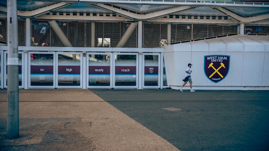 A person running past the exterior entrance of West Ham United's football stadium, visible with the club's blue and yellow crest on a white wall. The entrance features a large, modern glass and metal structure with sliding doors, displaying partially obscured text on a purple background, including the words 'so high nearly reach.' The pavement in front is a mix of light and dark textured surfaces, with a metal pole on the left side. The scene is outdoors during daytime with natural lighting, and the individual is wearing a light grey shirt, dark shorts, a face mask, and sneakers. This setting relates to house removals and relocation services, as it suggests transportation or moving activities near a well-known stadium, similar to connecting logistics and home relocation processes that companies like Man with Van West Ham may handle.
